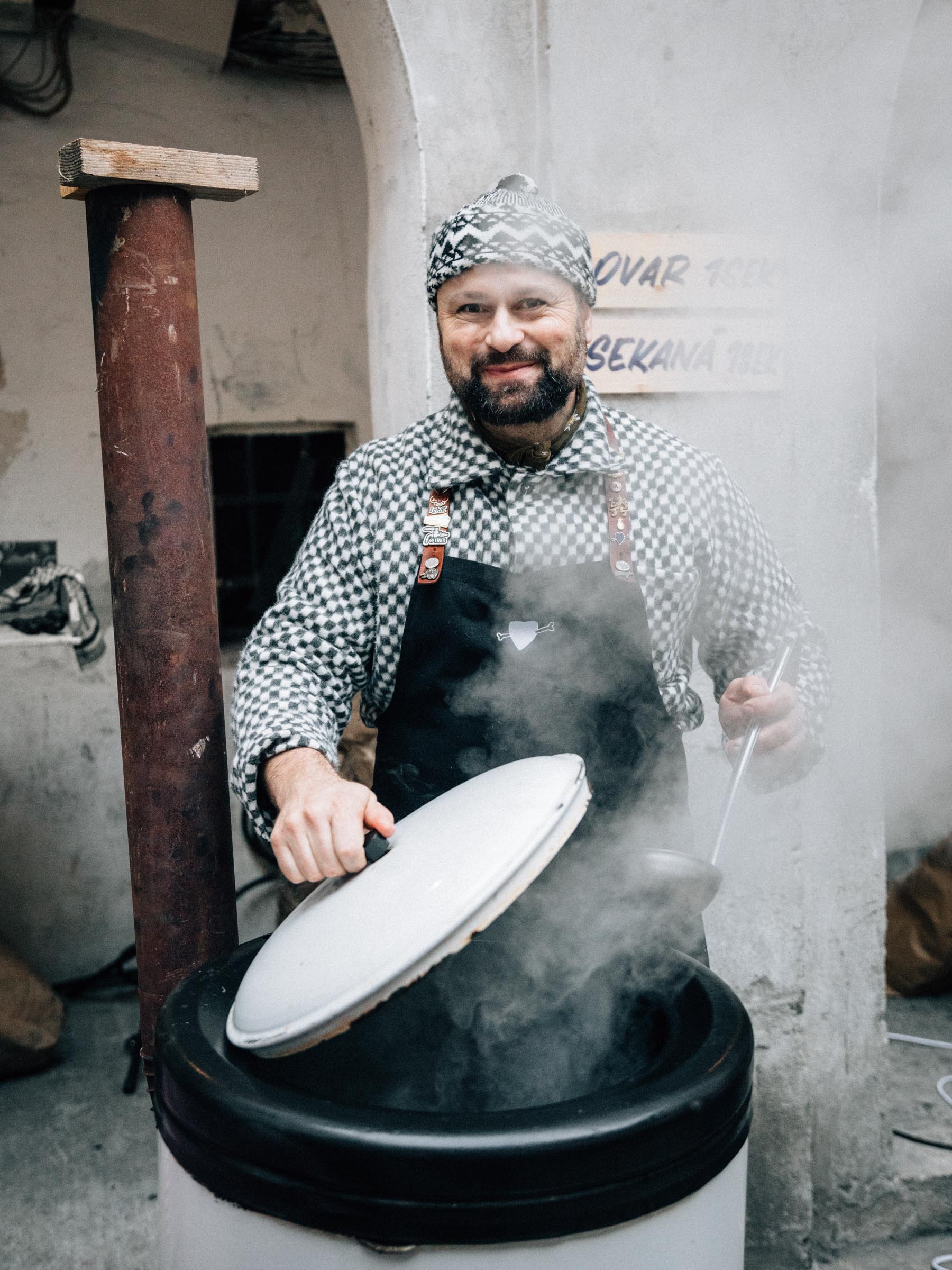 Muž v zástěře a kostkované košili s čepicí vaří u velkého hrnce, ze kterého stoupá pára.