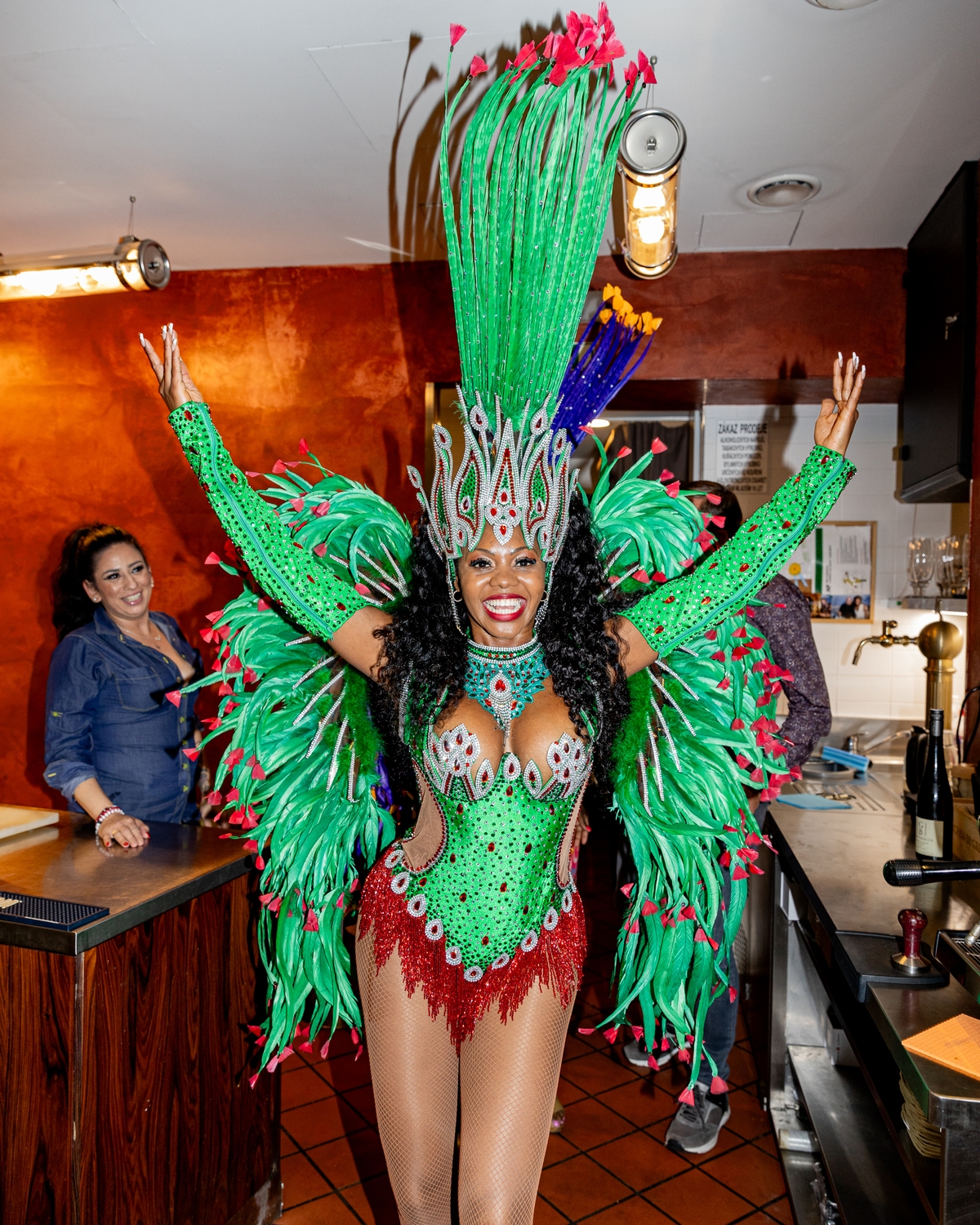 Tanečnice v pestrém zeleno-červeném karnevalovém kostýmu s bohatou čelenkou a pery pózuje s rukama nahoře v restauraci Brasileiro U Zelené žáby.