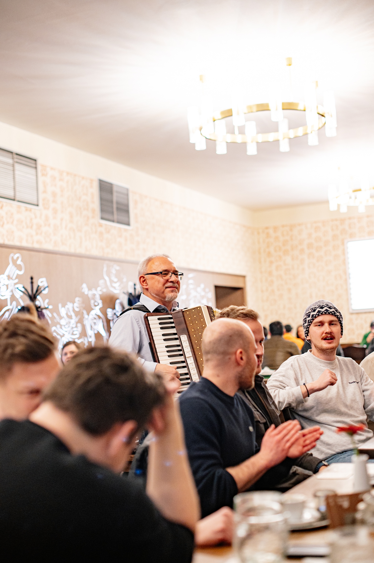 Muž hrající na akordeon mezi hosty v restauraci, lidé u stolu zpívají a tleskají.