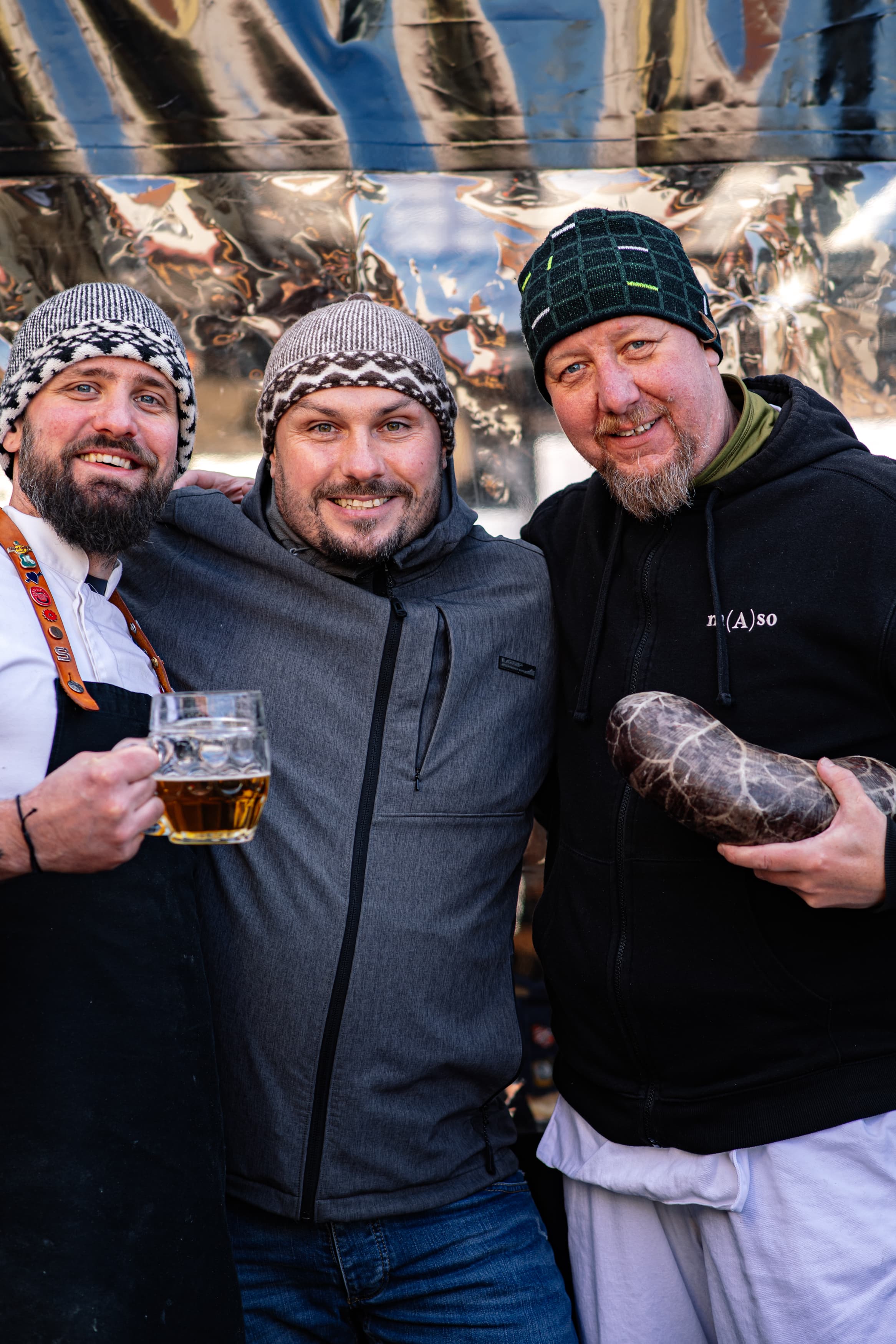 Tři muži v zimním oblečení se usmívají, jeden drží pivo a druhý tradiční zabijačkový výrobek.