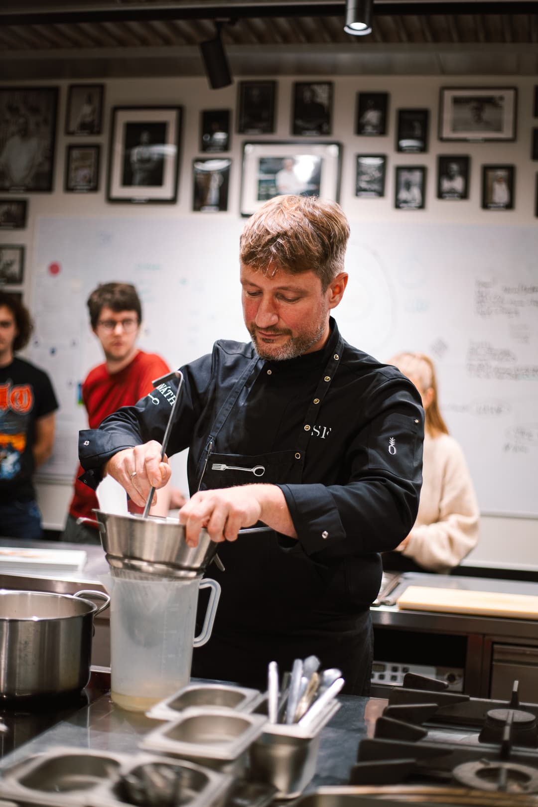 „Kuchař v černé uniformě přecedí omáčku přes síto během kulinářské prezentace, za ním sledují účastníci.