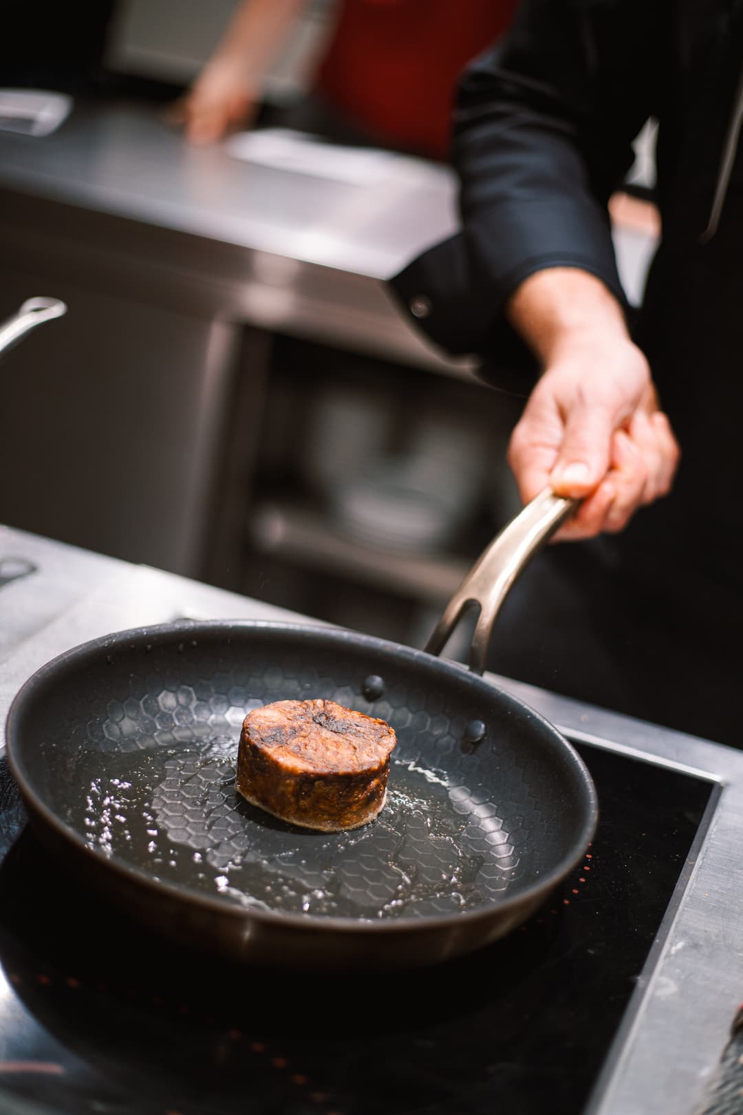 Kuchař držící pánev s opečeným kusem masa při přípravě pokrmu v profesionální kuchyni.