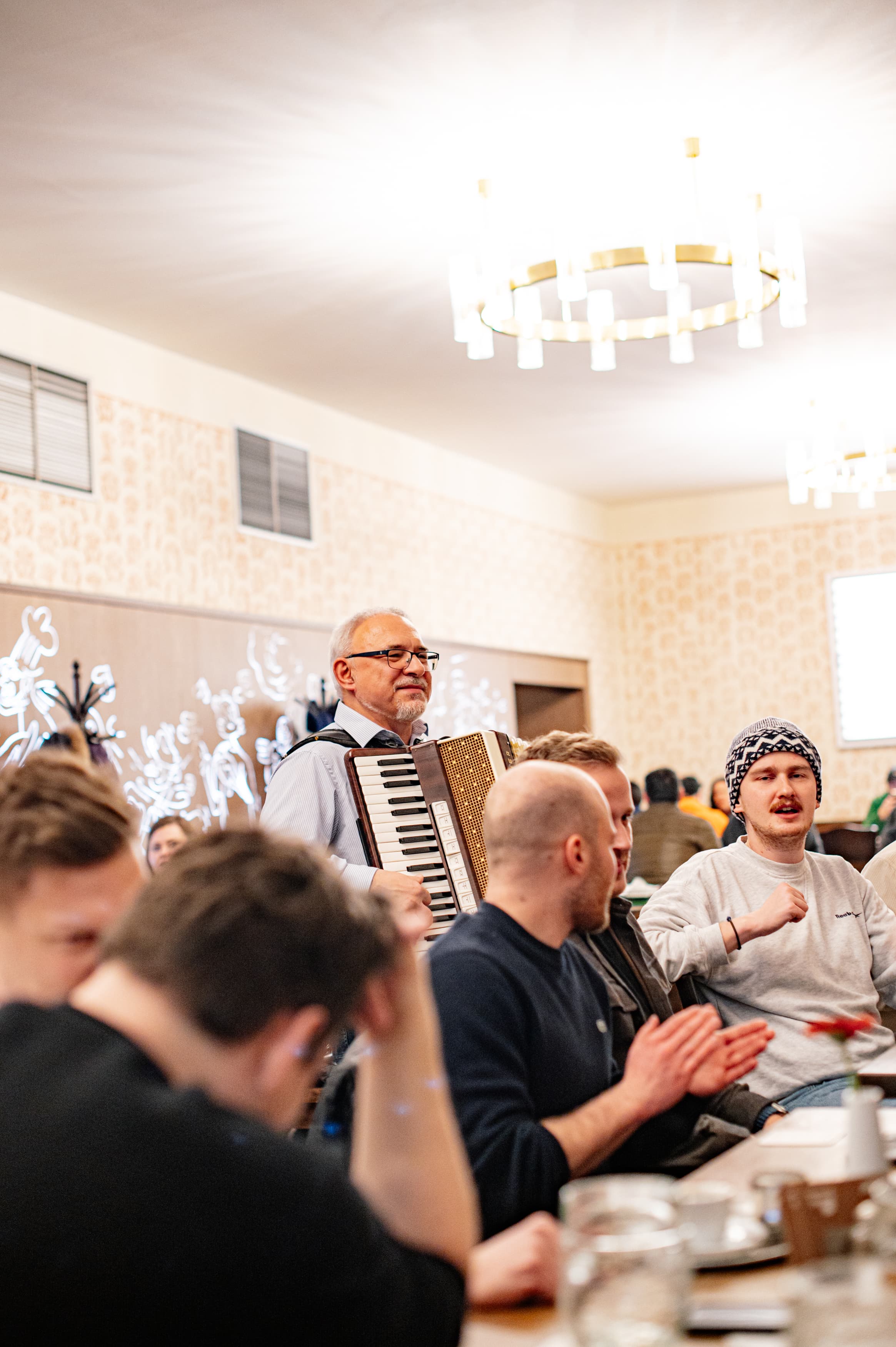 Muž hrající na akordeon mezi hosty v restauraci, lidé u stolu zpívají a tleskají.