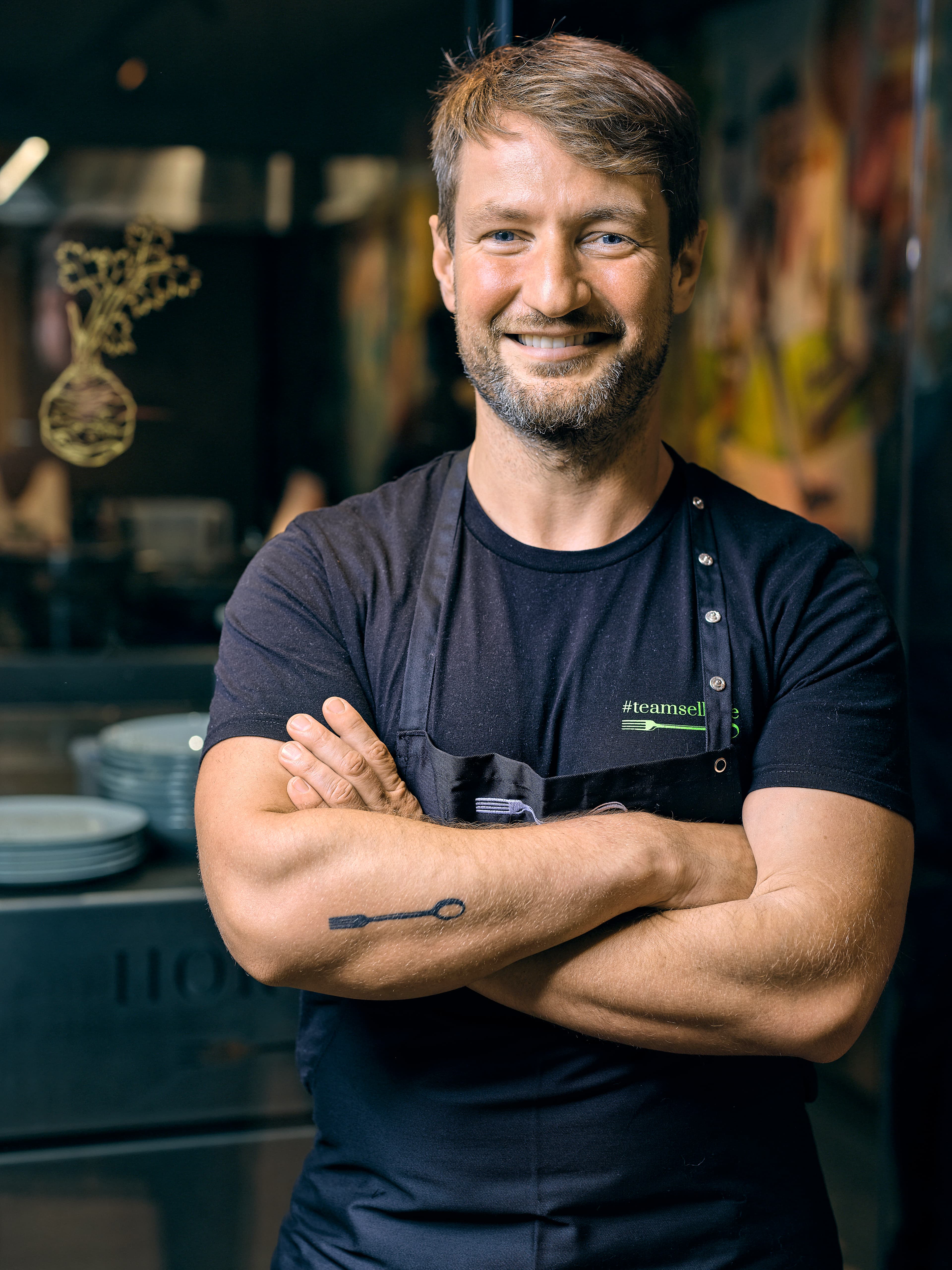 Portrét Sebastiana Franka, michelinského šéfkuchaře z berlínské restaurace Horváth, v kuchařské zástěře.  Sebastian Frank se usmívá a pózuje s překříženýma rukama, připravený na besedu v Ambiente.