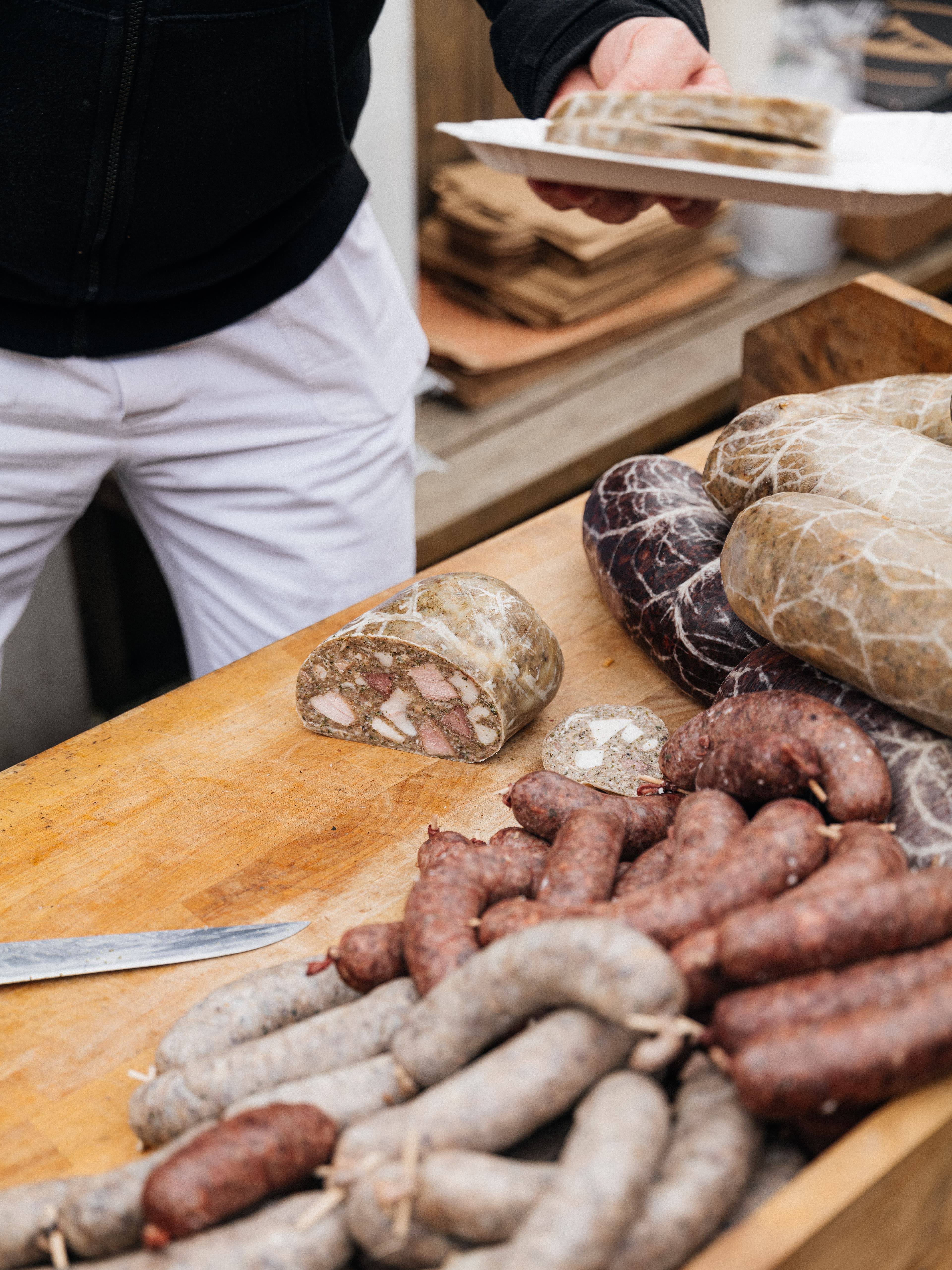 Detail stolu s domácími zabijačkovými výrobky, kuchař podává plátek tlačenky.