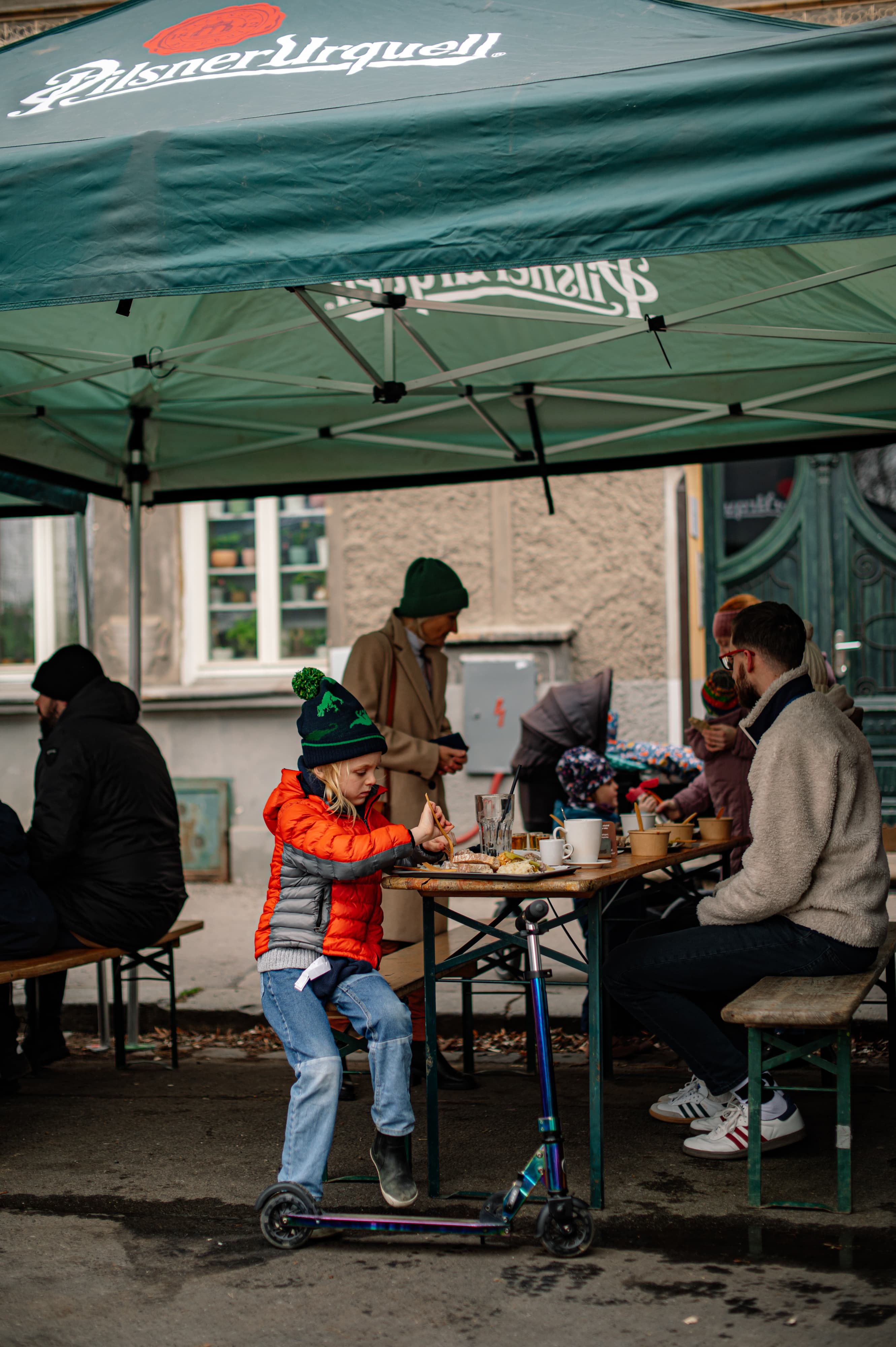 Rodiny sedící pod stánkem Pilsner Urquell, dítě u stolu s koloběžkou během venkovní akce v Lokálu Pod Divadlem.
