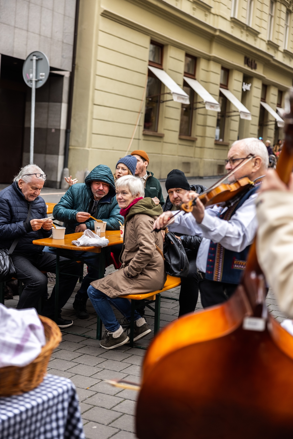 Skupina lidí sedící u venkovního stolu na ulici, poslouchající hudebníky hrající na housle a kontrabas.