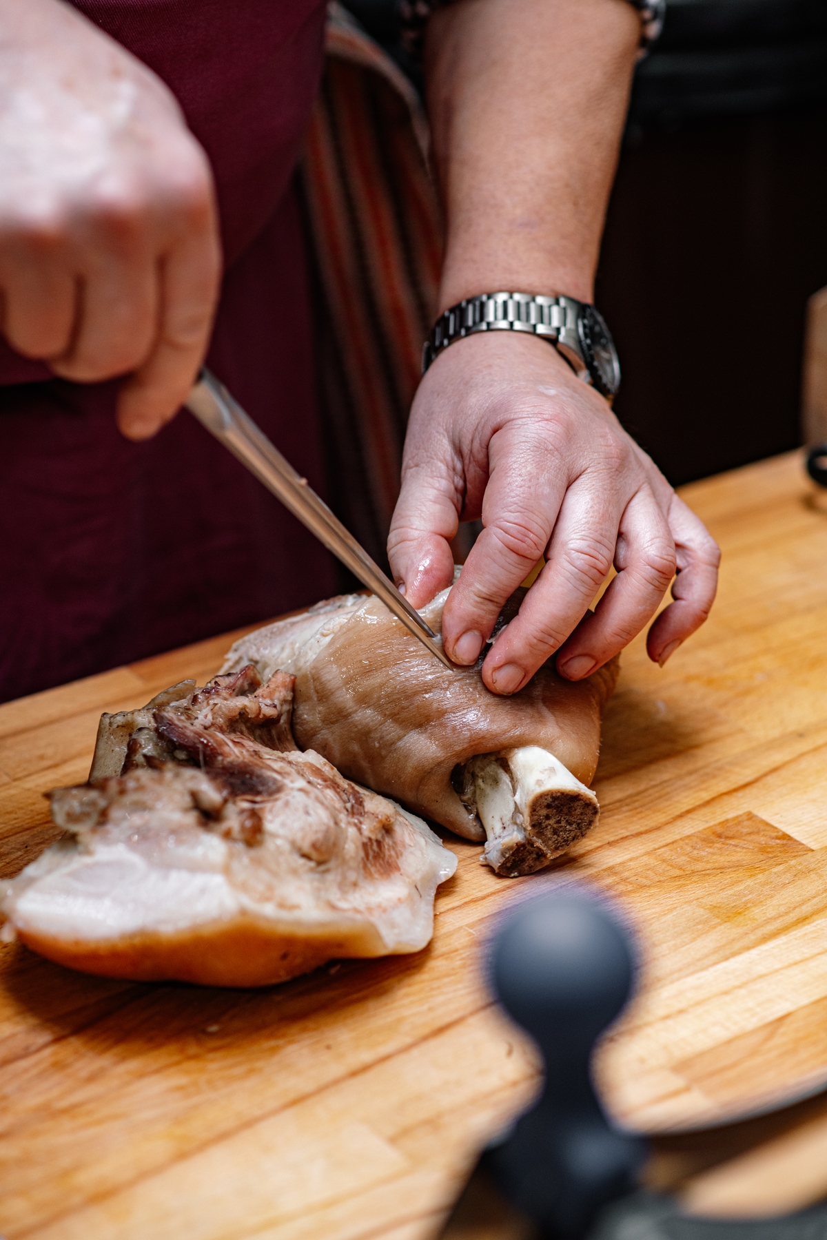 Osoba krájící vařenou vepřovou nohu na prkénku, detail rukou s nožem.
